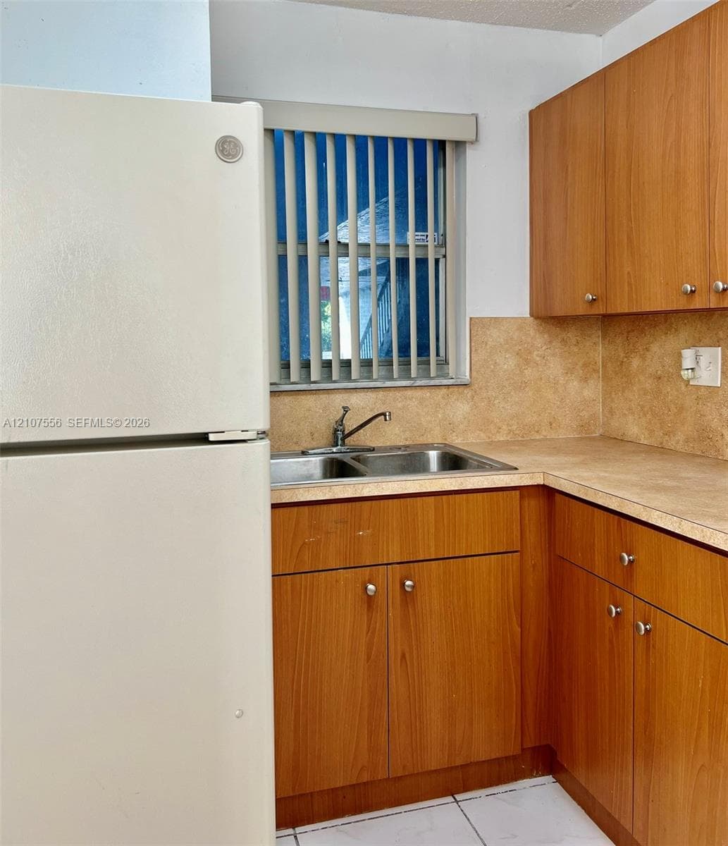 Kitchen featuring wood-finish cabinetry, neutral solid-surface countertops, stone-finish backsplash, stainless steel double-basin sink with faucet, and white tile flooring