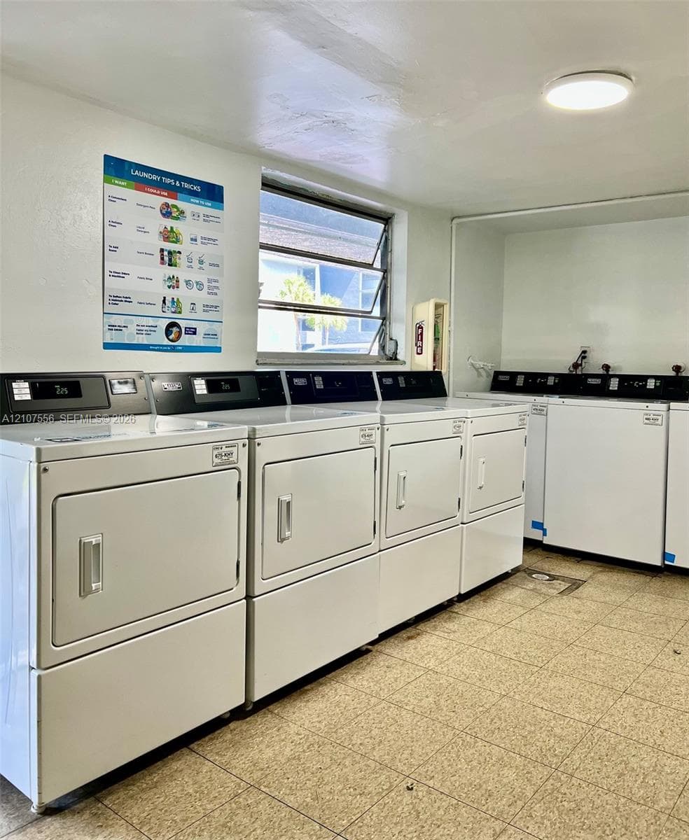 Utility room featuring multiple high-capacity white washing machines and dryers, ceramic tile flooring, a horizontal jalousie window, and overhead flush-mount lighting