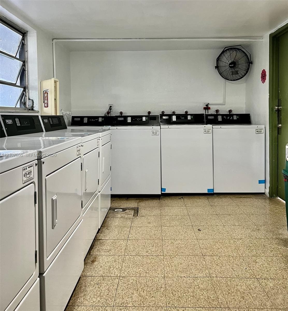 Utility room featuring four white washing machines, four white dryers, durable tile flooring, and a mounted wall fan