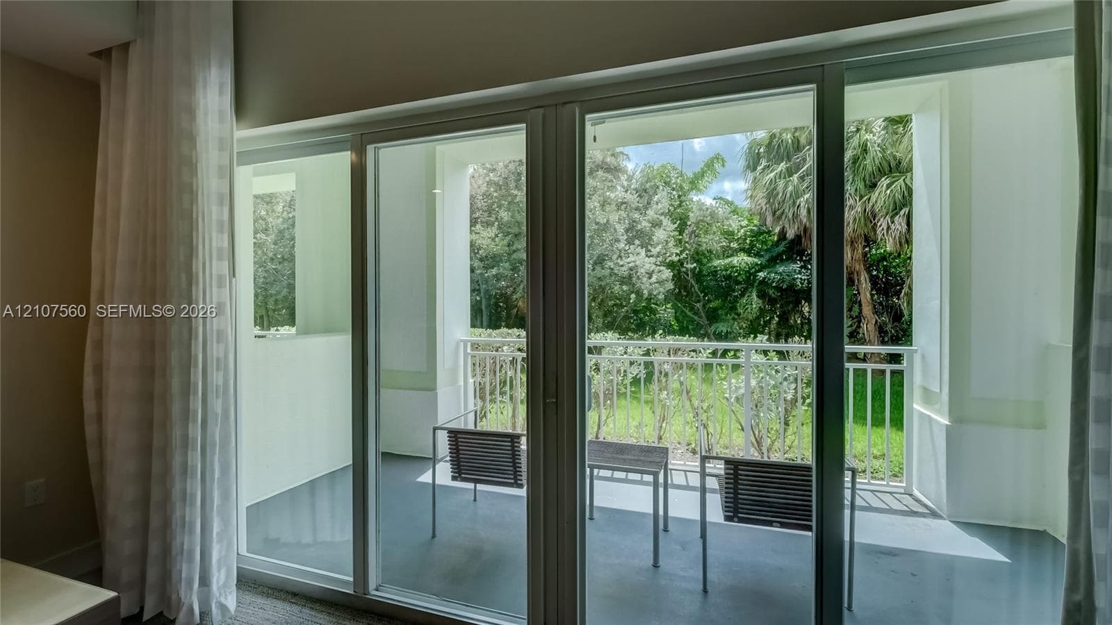 Floor-to-ceiling sliding glass doors opening to a covered balcony with white metal railings and greenery backdrop