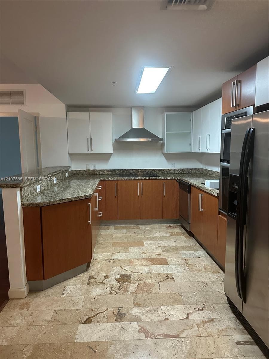 Kitchen featuring two-tone wood-finish and white cabinetry, stone-surface countertops, stone tile flooring, stainless steel appliances, and a wall-mounted chimney-style range hood
