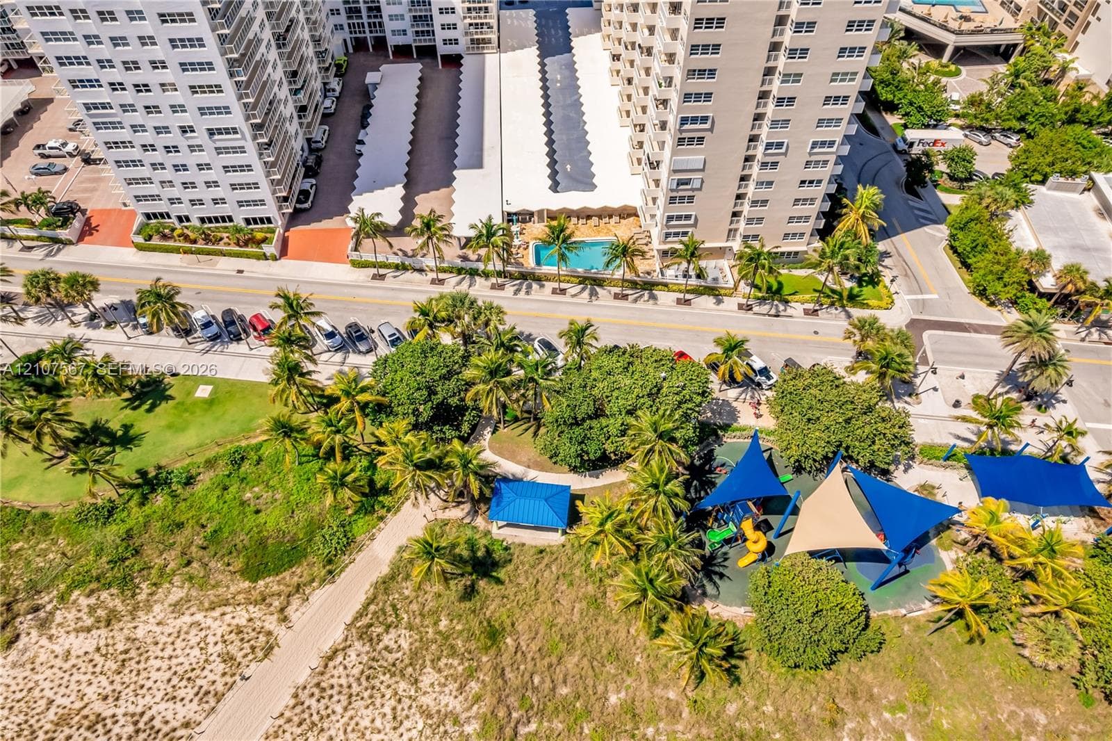 Aerial perspective featuring a high-rise residential structure with covered parking, a swimming pool, and proximity to a coastal park with shaded playground equipment and sandy terrain