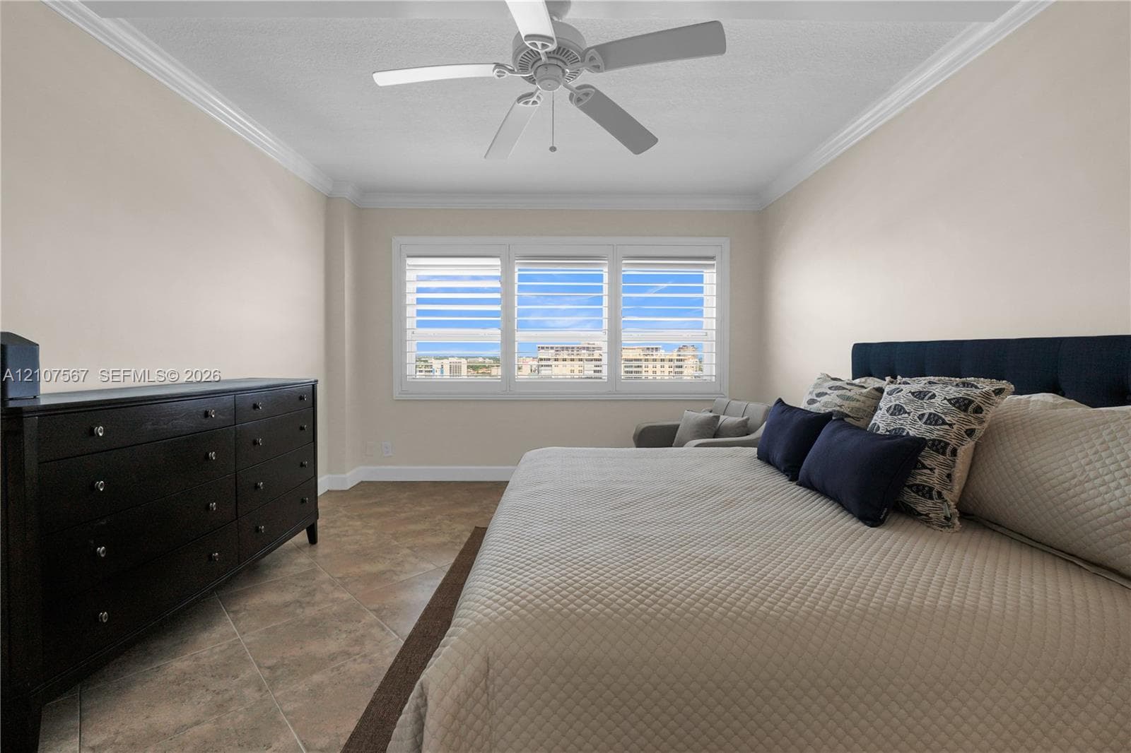 Neutral-toned interior featuring large-format tile flooring, multi-pane window configuration with plantation-style shutters, ceiling-mounted paddle fan, and crown molding