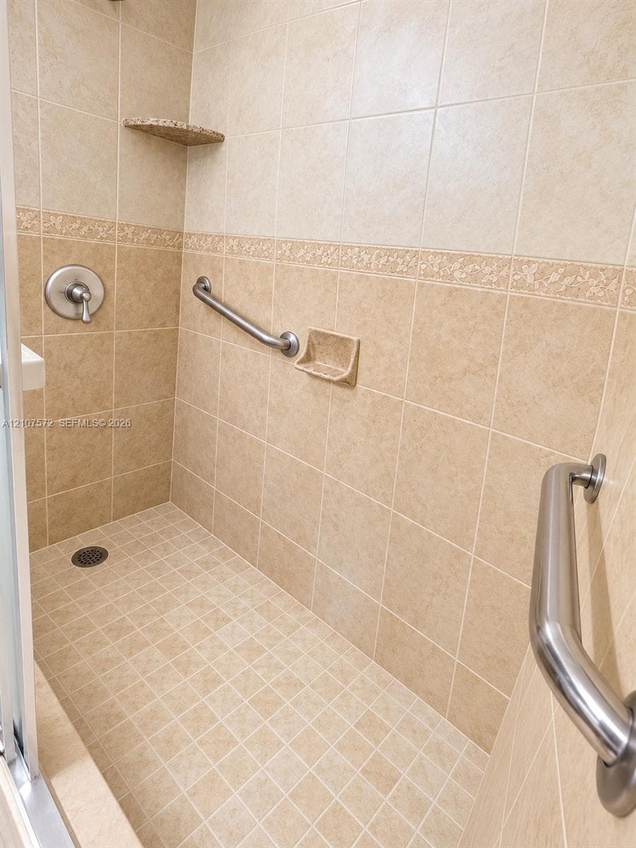 Walk-in shower featuring ceramic wall and floor tiling, decorative accent border, built-in corner shelf, and stainless steel grab bars