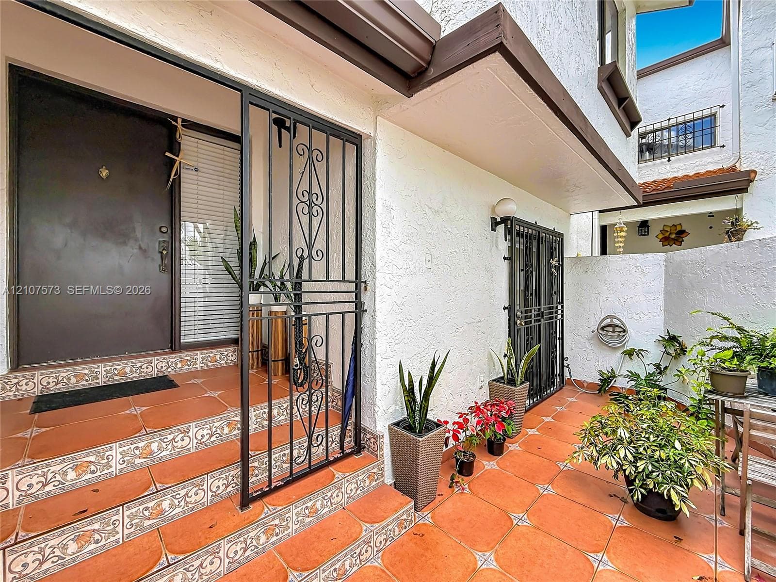 Private entryway featuring a decorative wrought iron security gate, textured exterior finish, and terracotta-style tile flooring with patterned accents