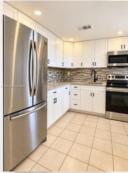 Kitchen featuring white shaker-style cabinetry, stainless steel appliances, multi-tonal linear tile backsplash, light-toned stone countertops, and tile flooring