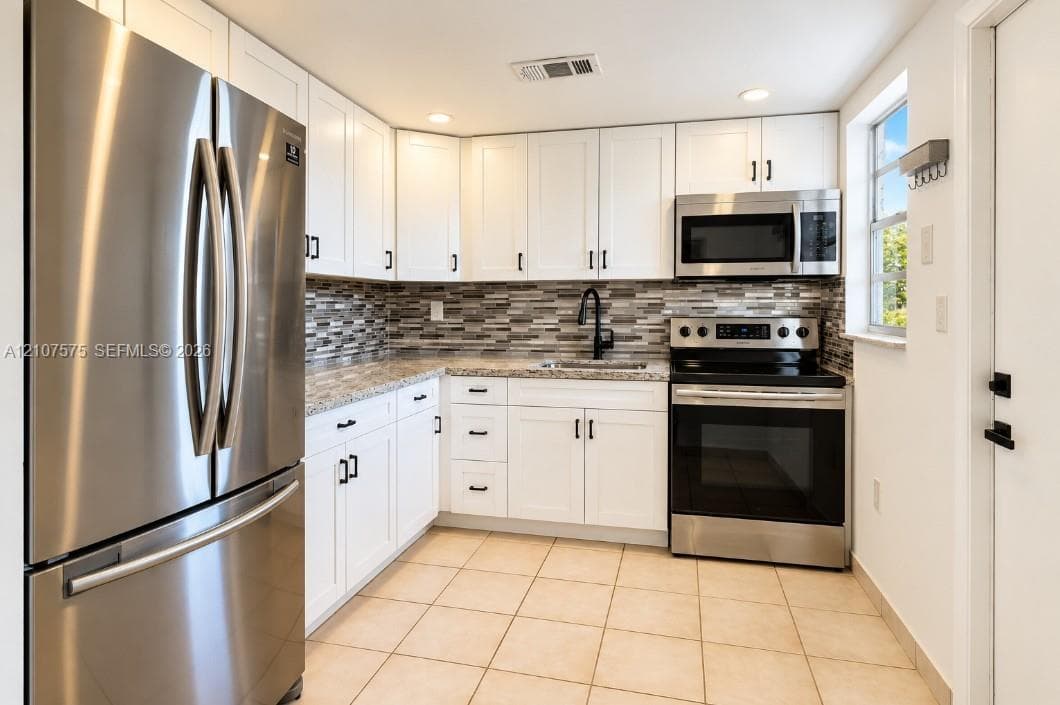 Kitchen featuring white cabinetry, stone-finish countertops, a multi-tonal tile backsplash, stainless steel appliances, and tile flooring