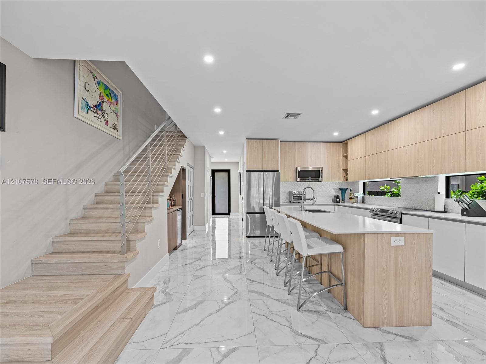 Modern kitchen featuring light wood-finish cabinetry, a large central island with solid-surface countertops, polished stone-finish floor tiling, and a custom staircase with a sleek horizontal railing