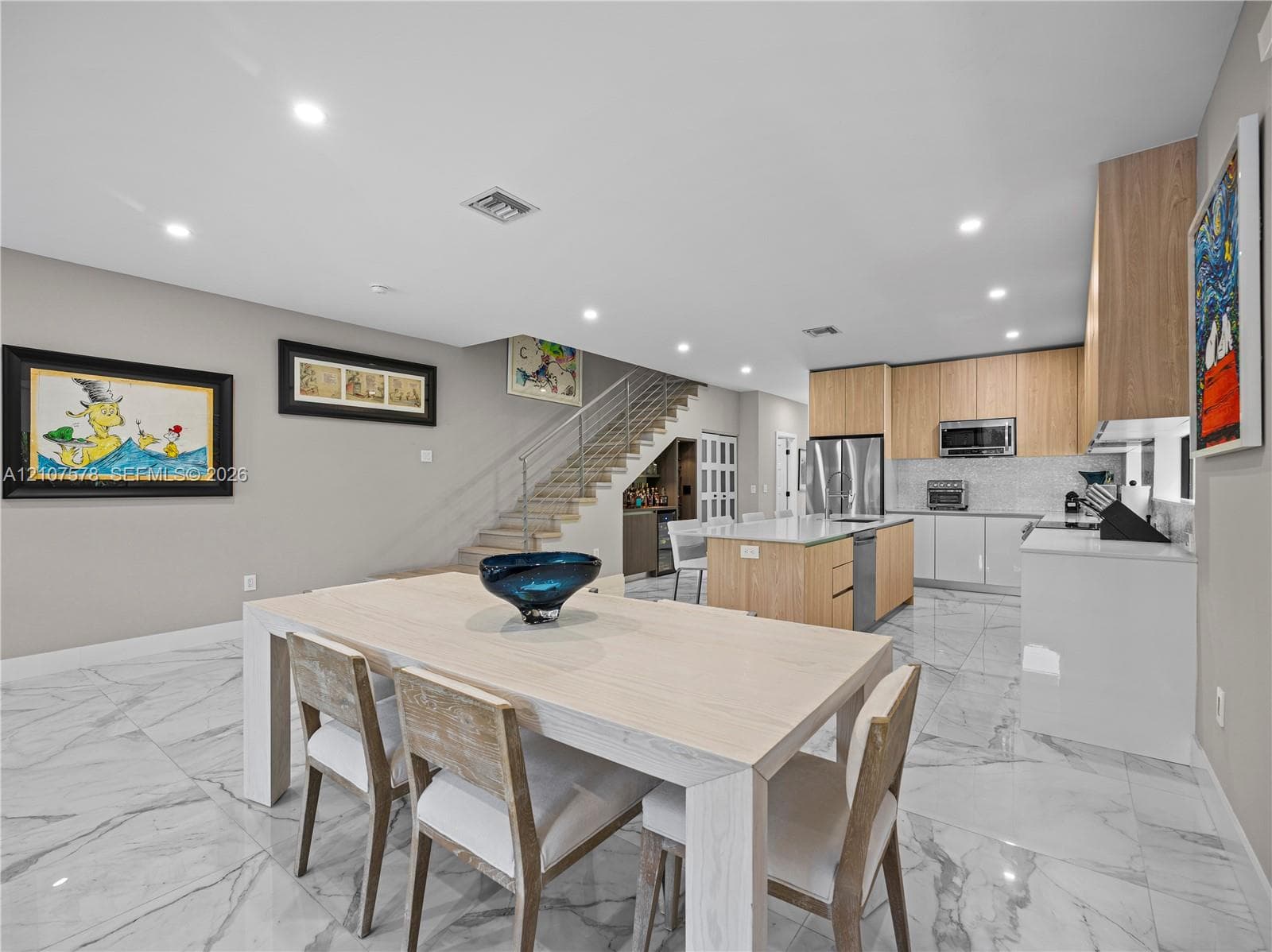 Modern kitchen featuring flat-panel wood cabinetry, a central island with white stone countertops, built-in stainless steel appliances, marble-patterned tile flooring, and a floating staircase with a metal railing