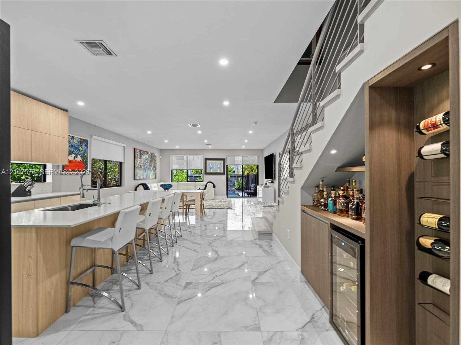 Open-concept kitchen featuring light wood-finish cabinetry, stone-surface countertops, large-format stone-finish flooring, a built-in wine cooler with integrated wine rack, and a staircase with metal railing