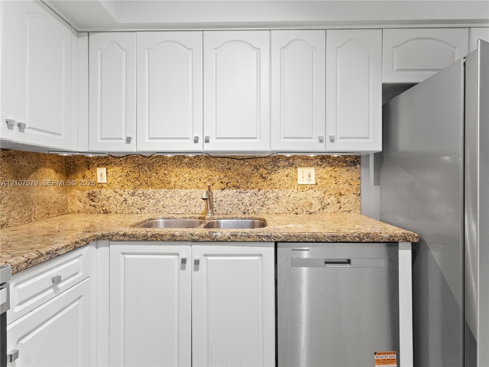 Kitchen featuring white cabinetry, stone-surface countertops, full-height stone backsplash, stainless steel dishwasher, and under-cabinet lighting