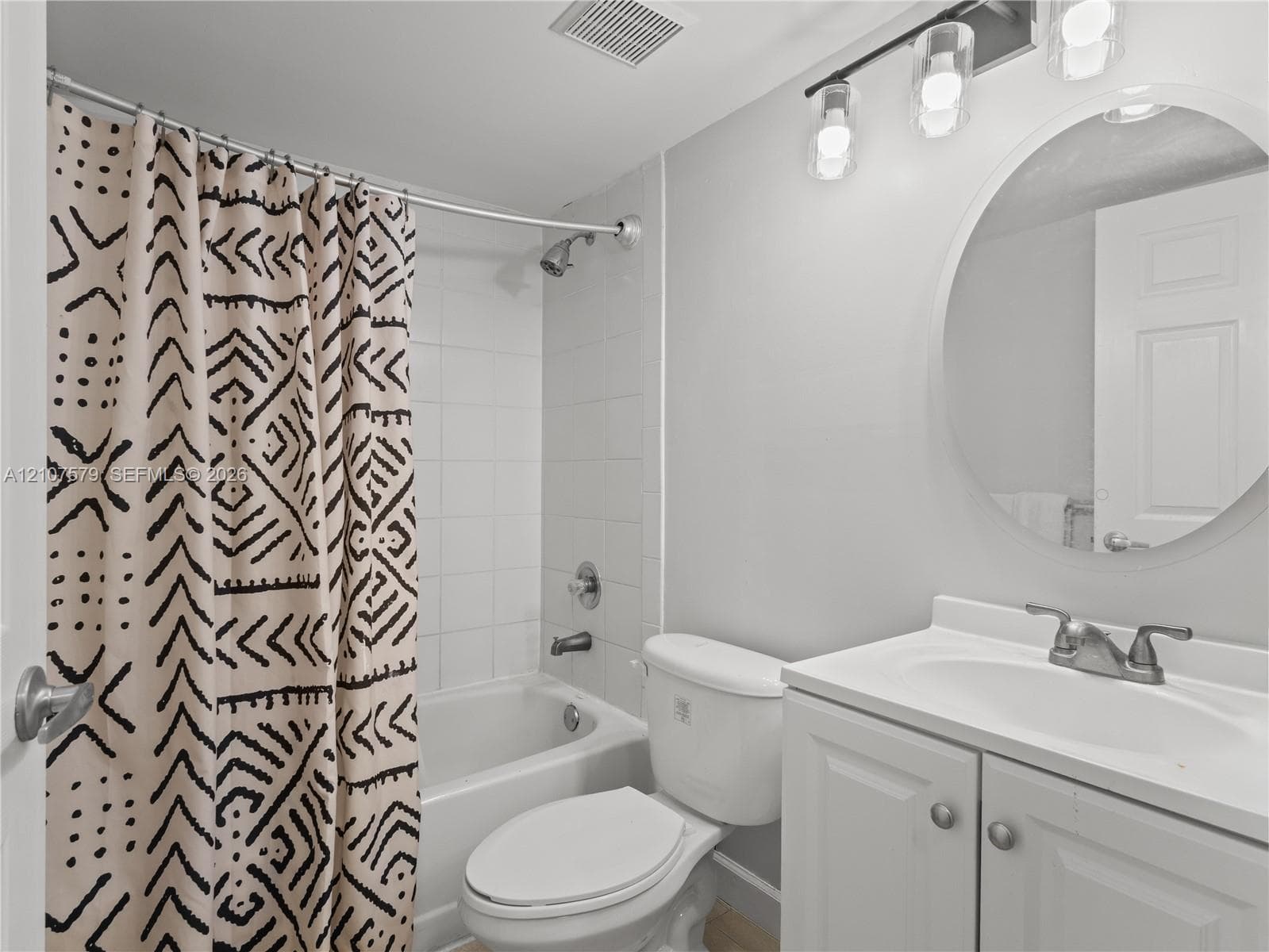 Bathroom featuring a vanity with integrated sink, circular mirror, three-light fixture, and a white tile surround shower-tub combination