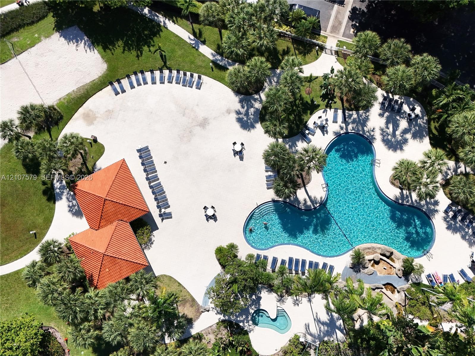 Community swimming pool featuring a large free-form basin, a secondary smaller pool, dual roofed structures with tile accents, and landscaped grounds with mature palm trees