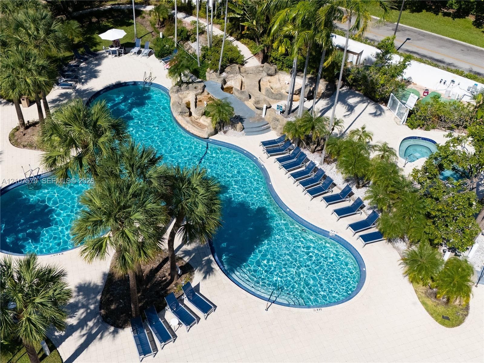 Curvilinear swimming pool with integrated rock waterfall feature, tiled patio decking, and separate elevated spa, surrounded by mature palm trees
