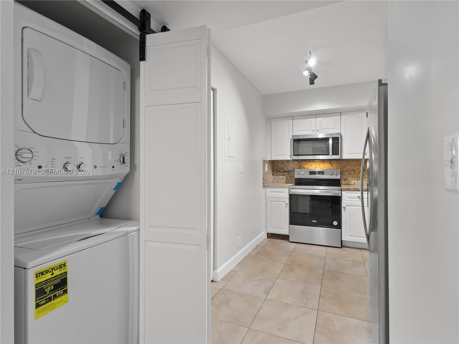 Laundry closet with stacked washer and dryer behind a sliding door, white shaker-style cabinetry, stainless steel appliances, stone-look tile flooring, and neutral stone-finish backsplash