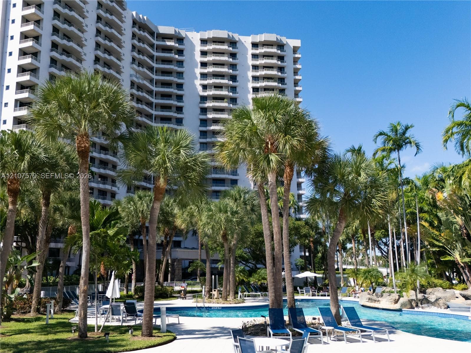 Multi-story high-rise building featuring private balconies, expansive swimming pool, hardscaped pool deck, and mature tropical landscaping