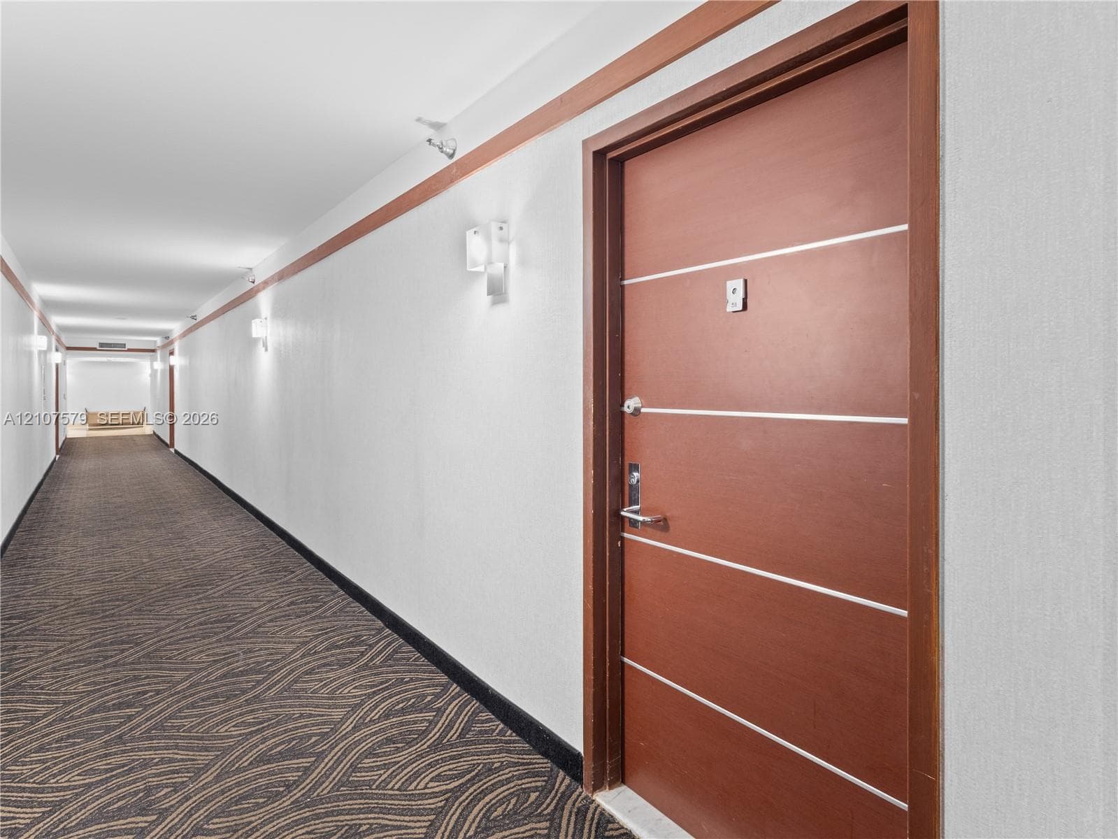 Interior hallway featuring a wood-finish door with horizontal metal inlay, patterned carpet flooring, wall-mounted sconce lighting, and decorative wood-trim wall accents
