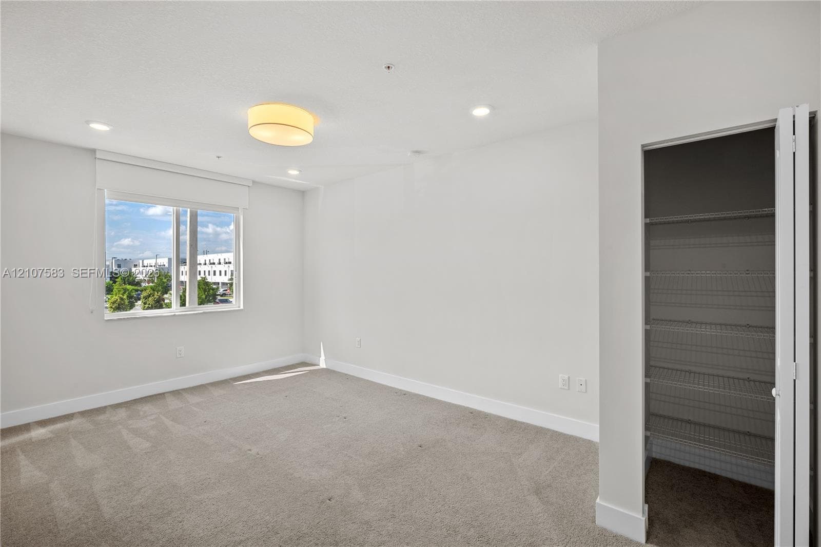 Neutral-toned interior featuring wall-to-wall carpeting, recessed lighting, a central drum light fixture, large window with roller shade, and a closet with white bi-fold doors and built-in wire shelving