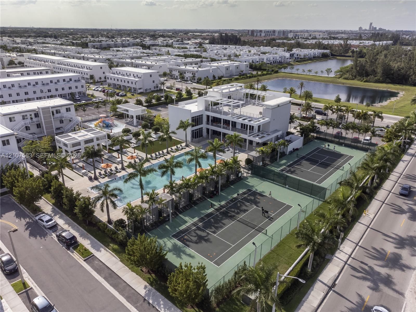 Aerial perspective of a community facility featuring a large swimming pool, two fenced tennis courts, and landscaped grounds with mature palm trees