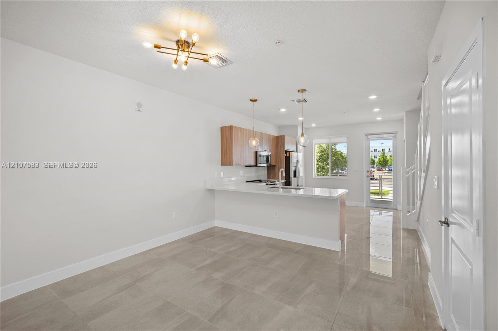 Open-concept interior featuring light-toned tile flooring, wood-finish cabinetry, a white countertop peninsula, and modern multi-light fixtures