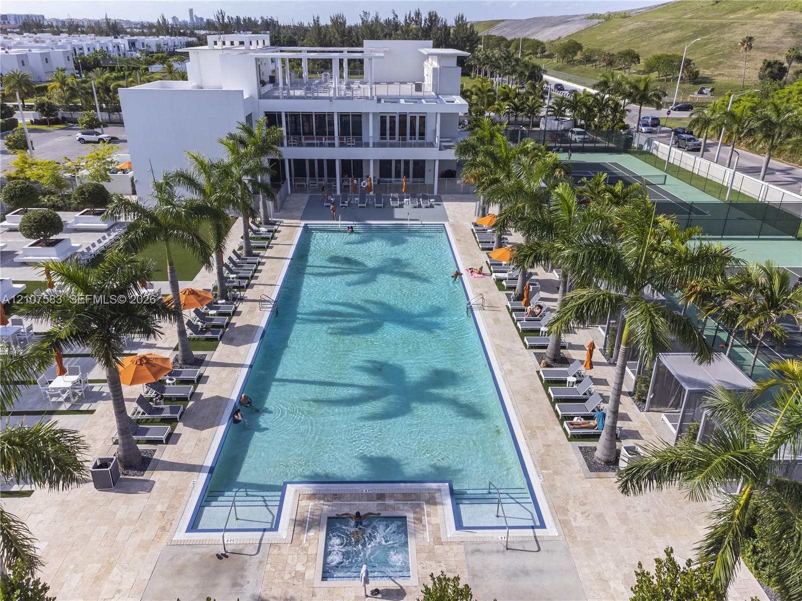 Rectangular swimming pool with integrated spa, stone-paver deck, professional-grade tennis court, and perimeter landscaping with palm trees
