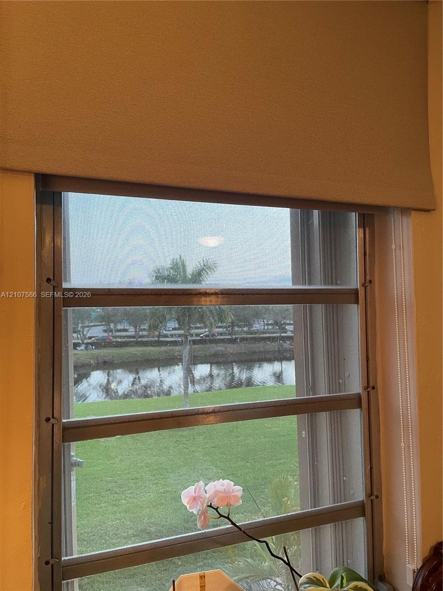 Multi-pane jalousie window featuring a neutral-toned roller shade and exterior views of a waterway, manicured lawn, and mature palm trees