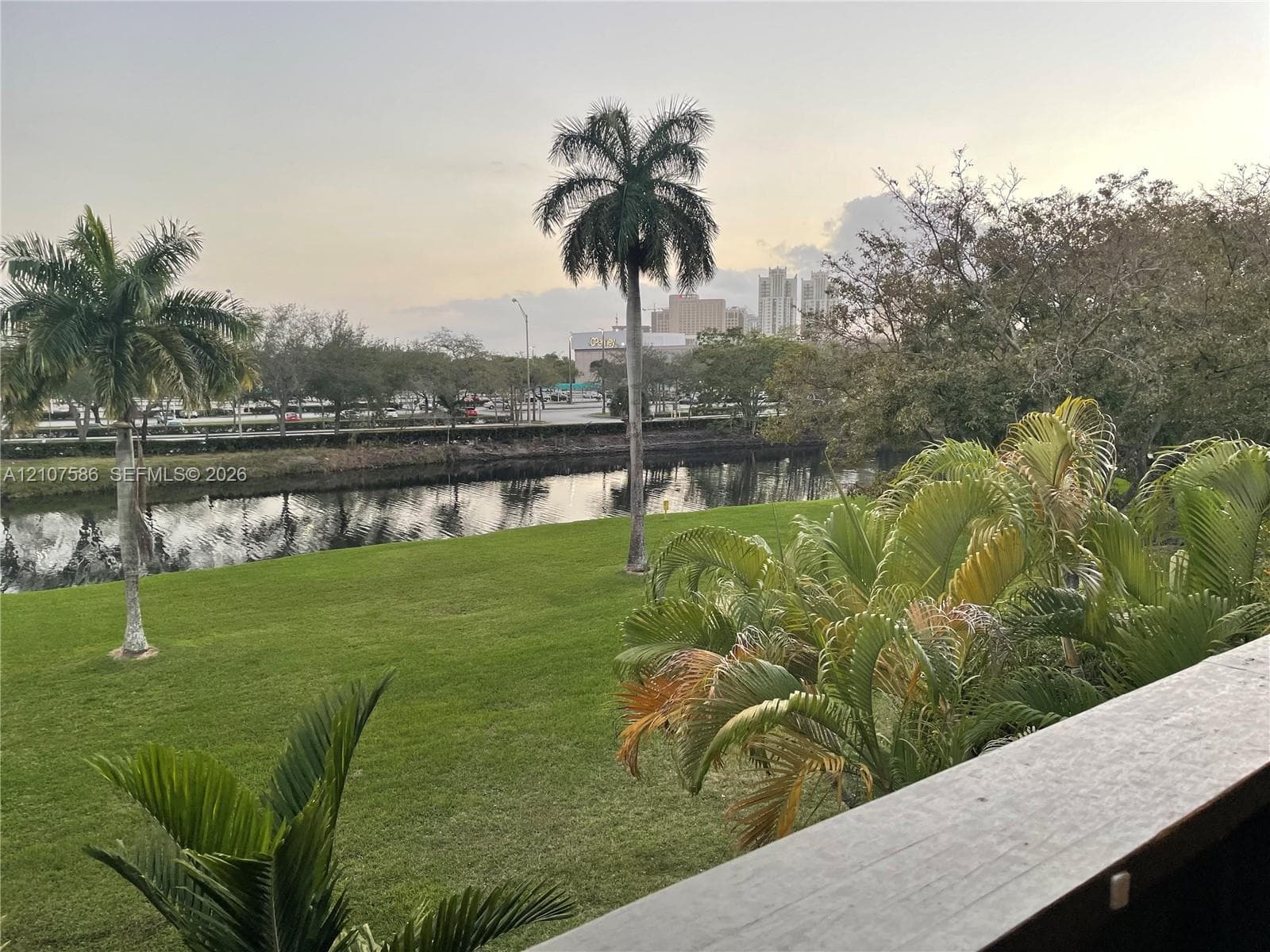 Outdoor balcony featuring a wood-finish railing overlooking a manicured lawn, a canal, and mature palm trees
