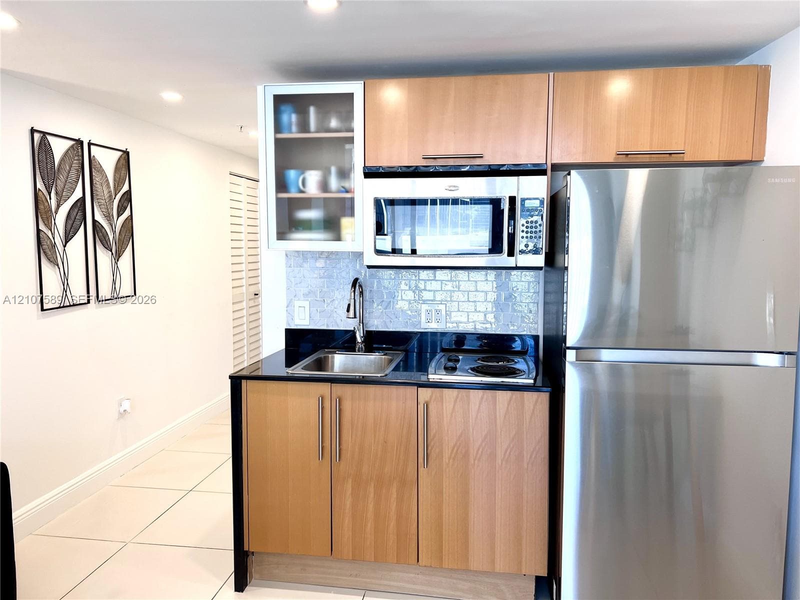 Kitchenette featuring flat-panel wood-finish cabinetry, black stone countertops, a stainless steel refrigerator, built-in microwave, and a glass-front upper cabinet