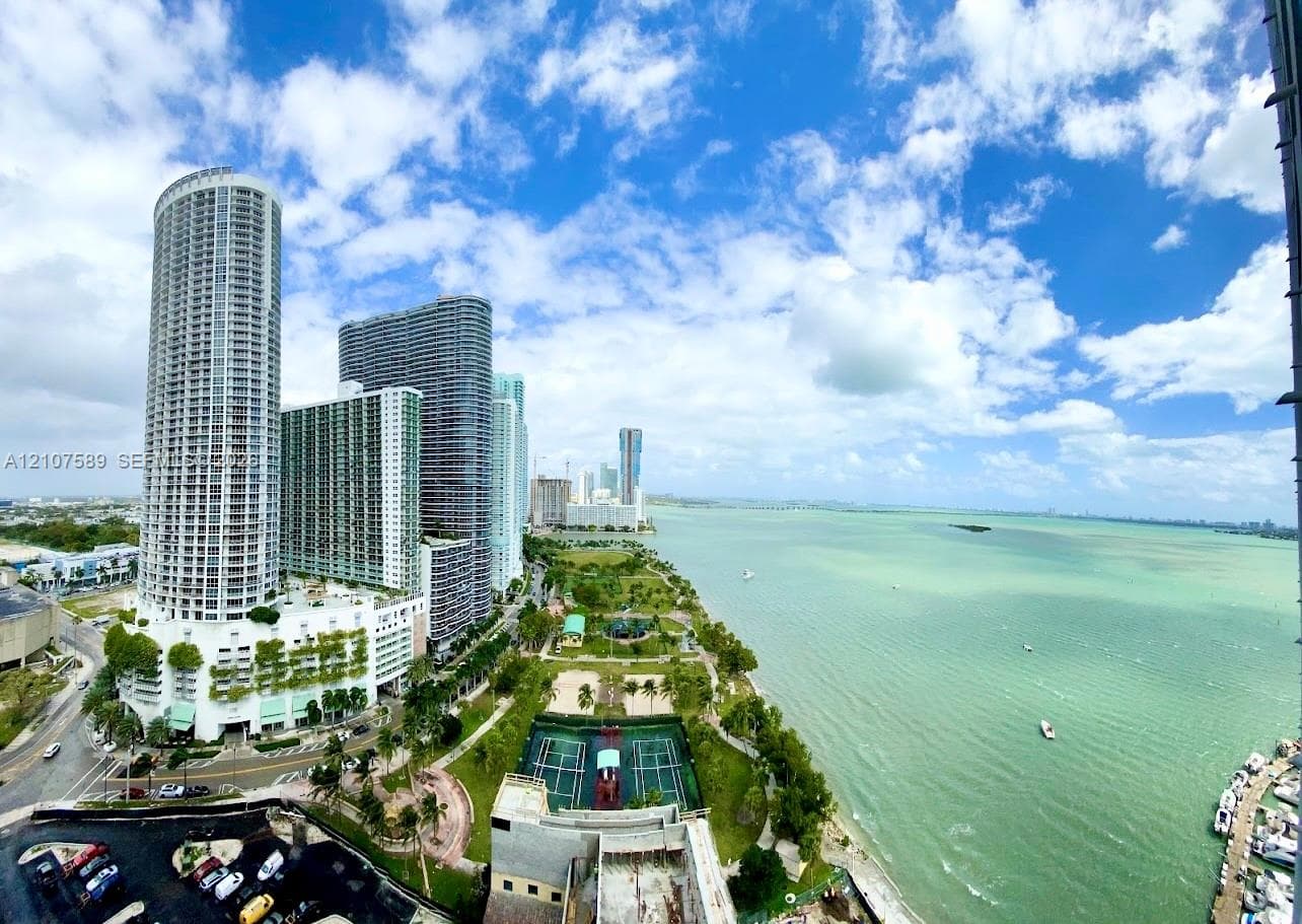 Panoramic coastal scenery featuring open water, a shoreline park, tennis courts, and high-rise urban architecture