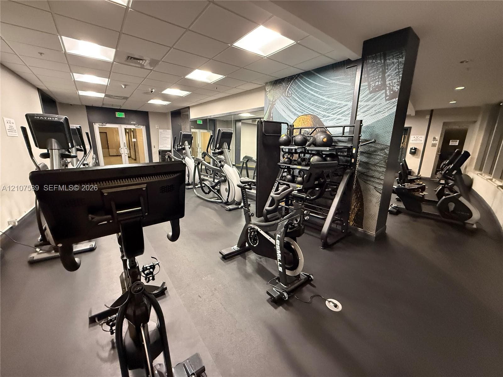 Fitness center featuring a drop ceiling, rubberized flooring, floor-to-ceiling mirror panels, and a decorative mural wall