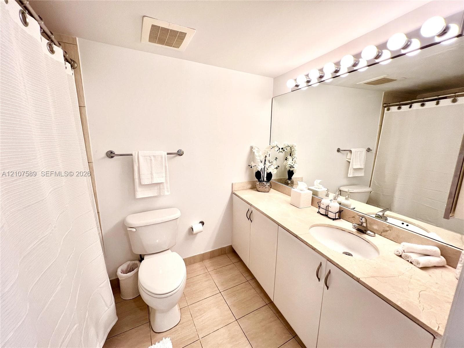 Bathroom featuring a full-width vanity with light-toned stone countertop, recessed oval sink, expansive wall-mounted mirror with integrated multi-bulb lighting, and tile flooring