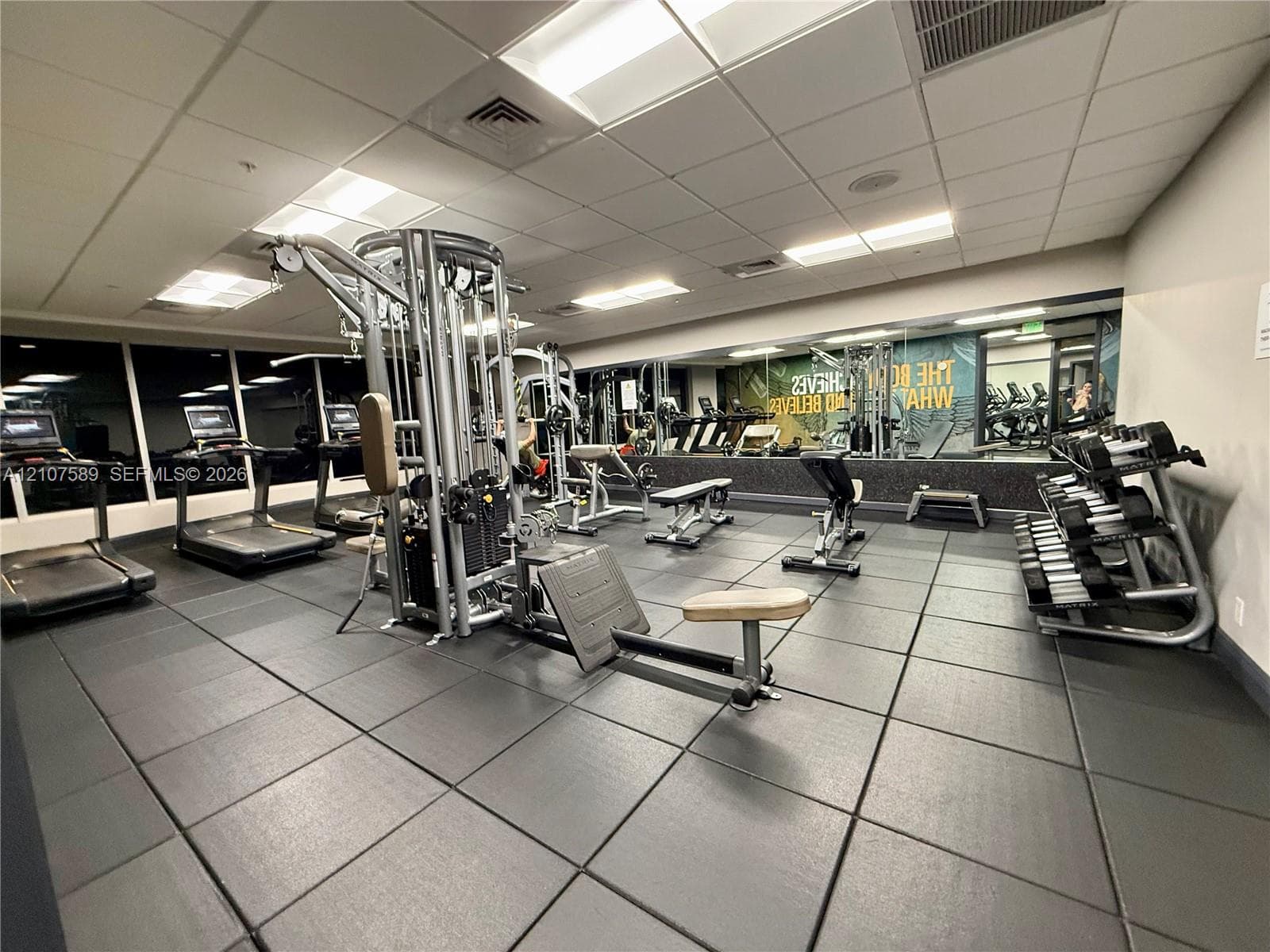 Fitness center featuring black rubberized flooring, a full-length wall mirror, drop ceiling with recessed lighting, and wall-to-wall windows