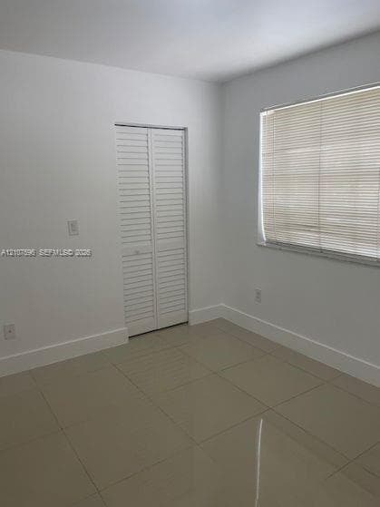 Neutral-toned tile flooring, white walls, and a closet with white louvered bi-fold doors, complemented by a window with installed horizontal blinds
