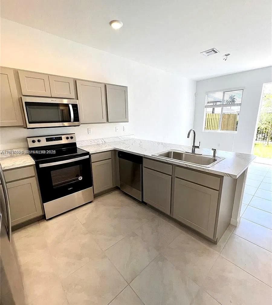 Kitchen featuring neutral-toned shaker cabinetry, stone-patterned countertops, stainless steel appliances, a dual-basin undermount sink with a gooseneck faucet, and large-format tile flooring