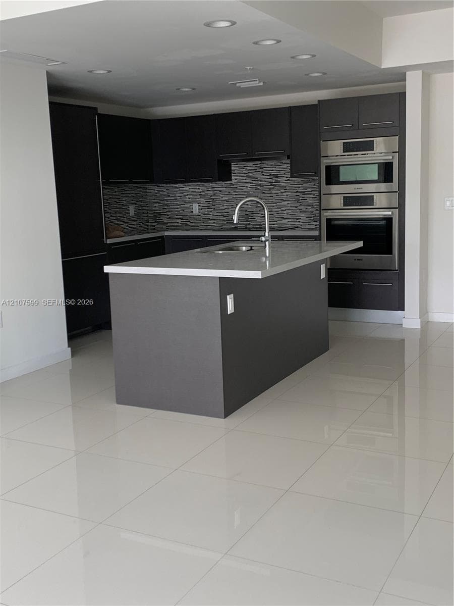 Kitchen featuring dark cabinetry, quartz countertops, a central island with a stainless steel faucet, stacked built-in stainless steel ovens, and large-format tile flooring