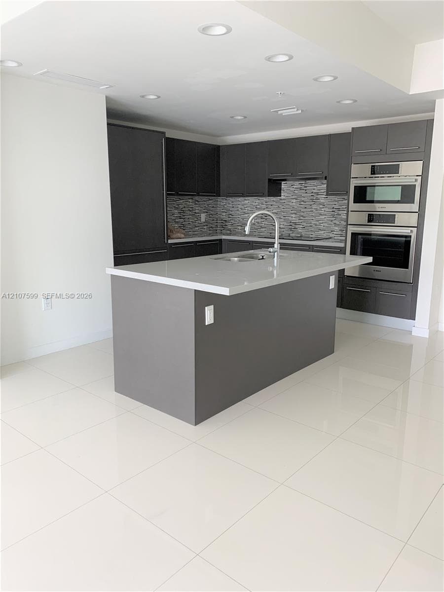 Kitchen featuring dark cabinetry, stone-finish countertops, a central island with an integrated sink, built-in stainless steel double ovens, and polished tile flooring