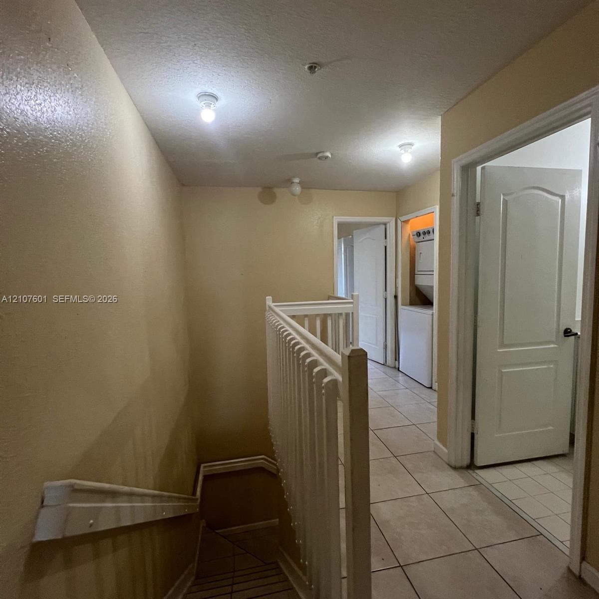 Staircase featuring a white baluster railing and handrail, neutral ceramic tile flooring, and integrated laundry closet with stacked appliance hookups