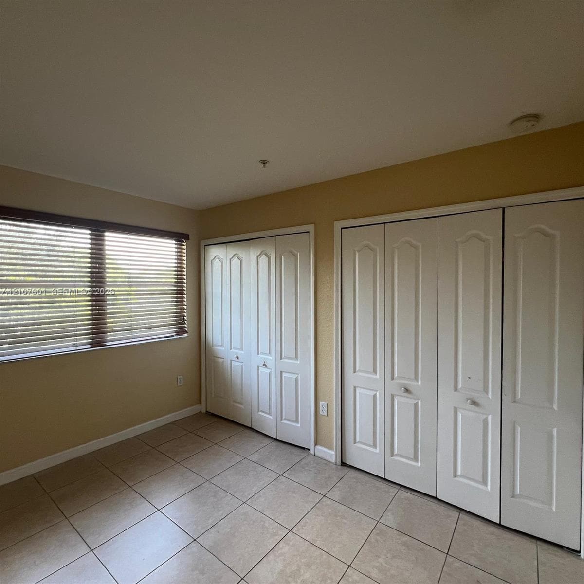 Interior room featuring tile flooring, two sets of white paneled bi-fold closet doors, and a window equipped with horizontal blinds