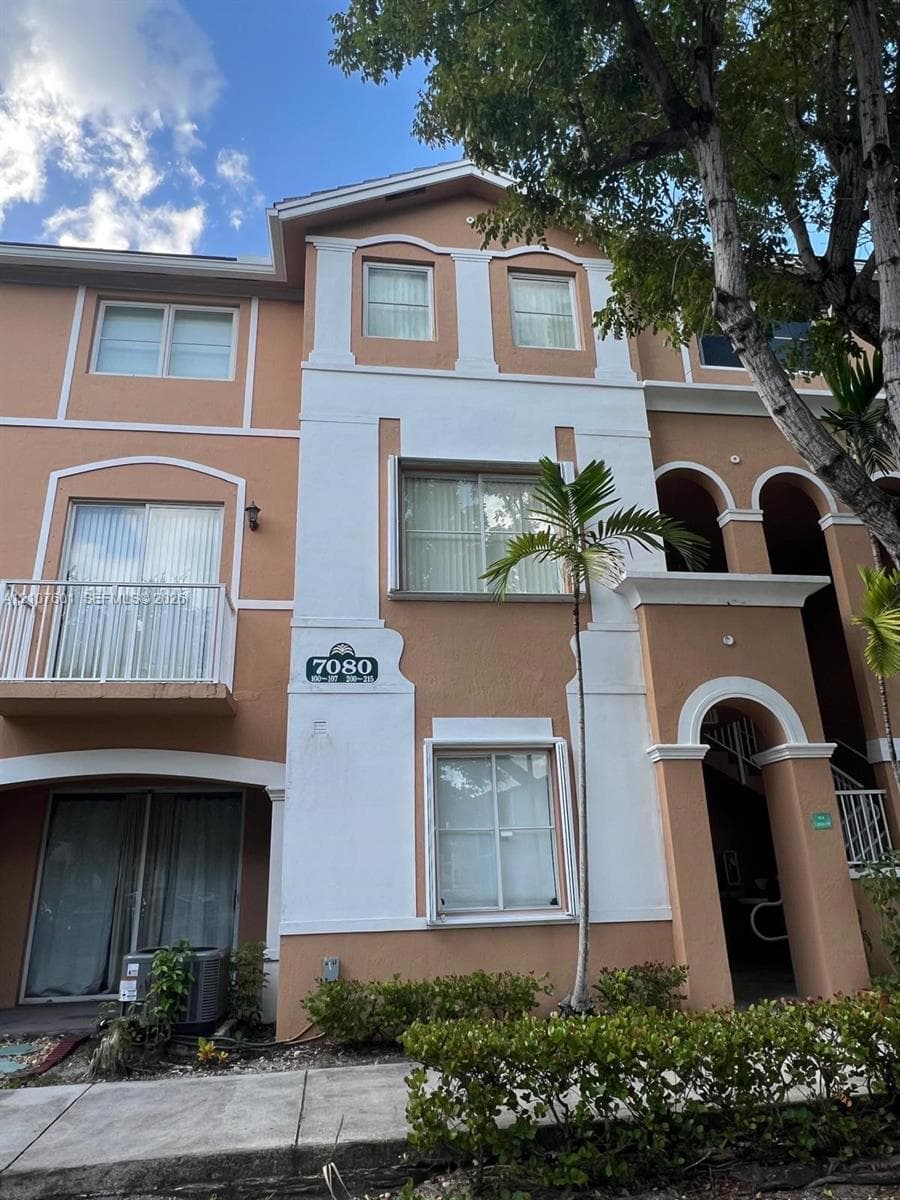 Multi-story building exterior featuring stucco cladding, arched architectural accents, a private balcony with metal railings, and a covered ground-level entry