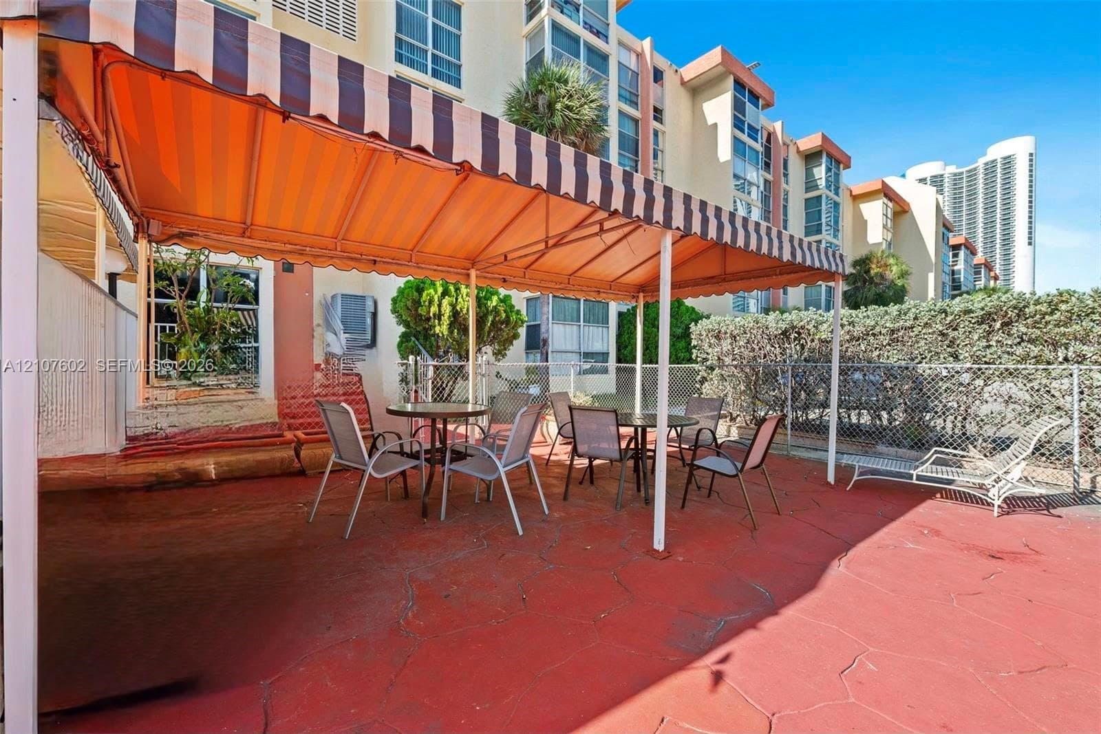 Ground-level patio featuring red-toned concrete flooring, a retractable striped fabric awning, and perimeter chain-link fencing