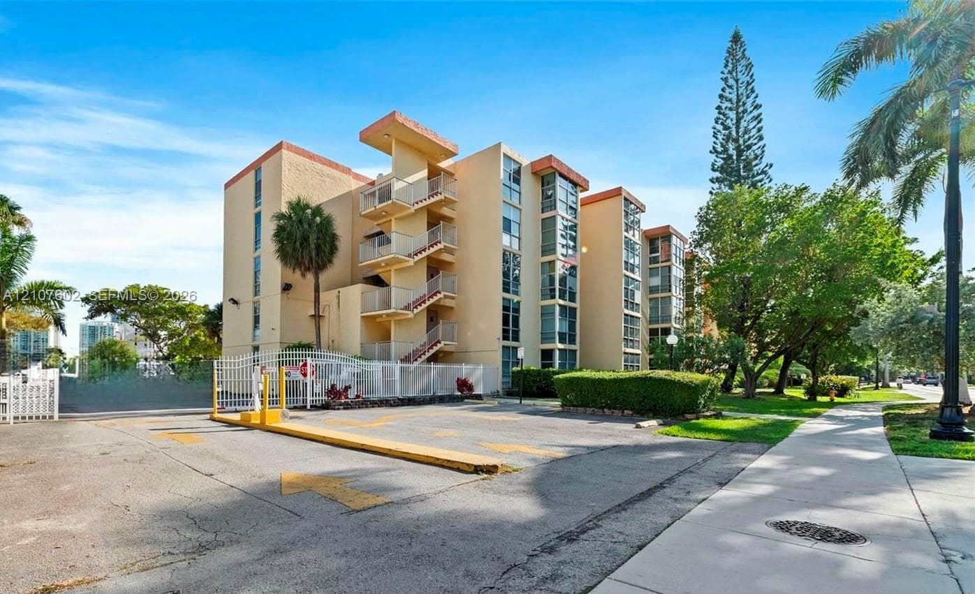 Multi-story residential building featuring a stucco exterior, integrated balconies, vertical window panels, and a gated parking area