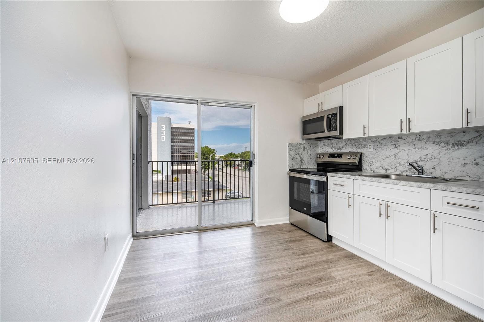 Kitchen featuring white shaker-style cabinetry, stone-patterned backsplash, stainless steel appliances, wood-finish flooring, and a sliding glass door opening to a private balcony