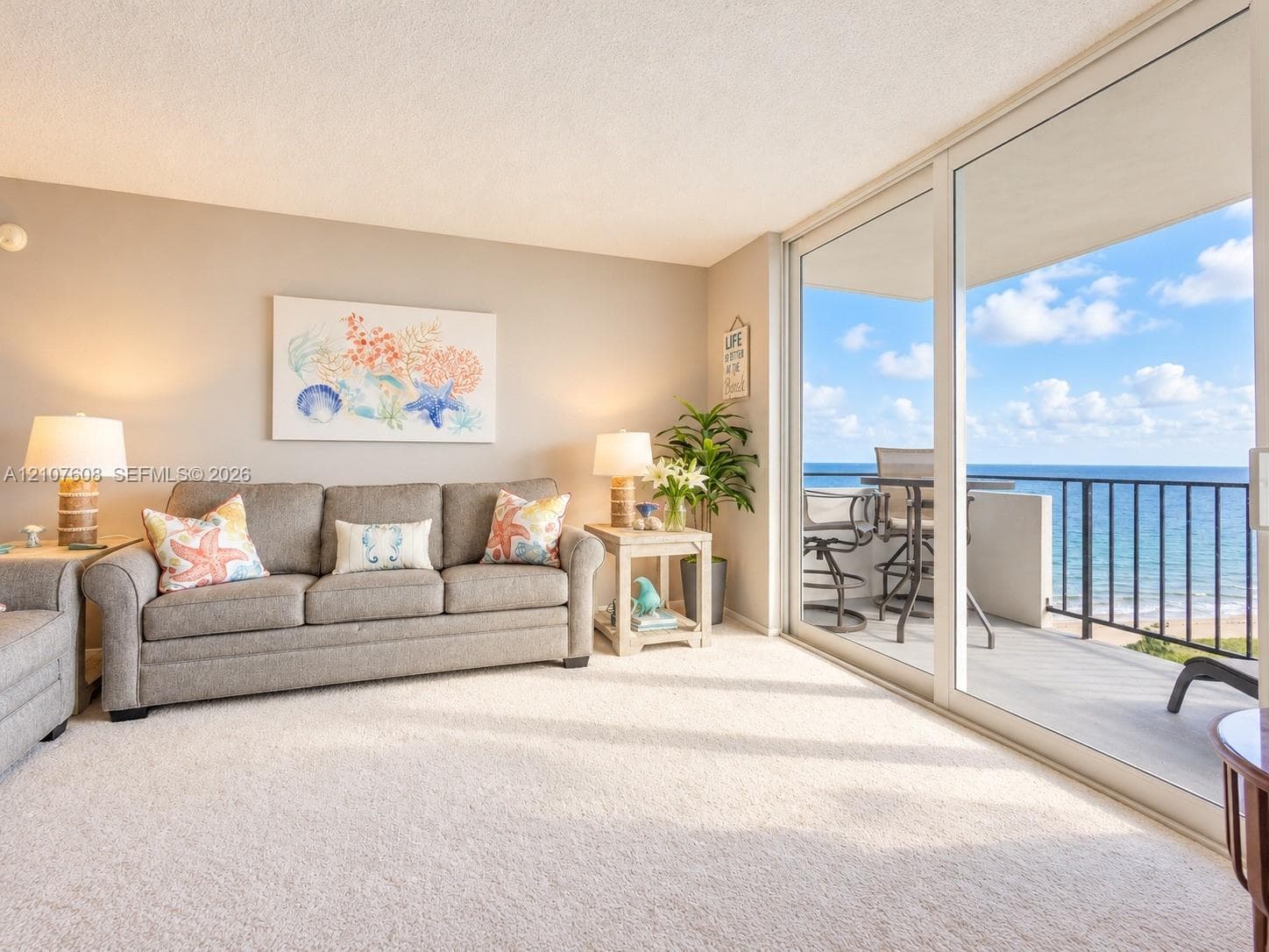 Living area featuring wall-to-wall carpeting and floor-to-ceiling sliding glass doors opening to a balcony with metal railings and coastal surroundings