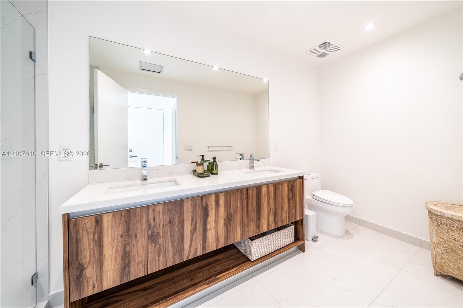 Dual-sink floating vanity with wood-finish cabinetry, white countertop, large wall-to-wall mirror, and glass-enclosed shower