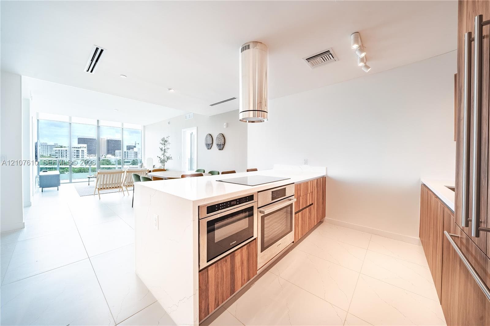 Modern kitchen featuring wood-finish cabinetry, white countertop surfaces, a stainless steel cylindrical range hood, built-in stainless steel double ovens, and large-format tile flooring