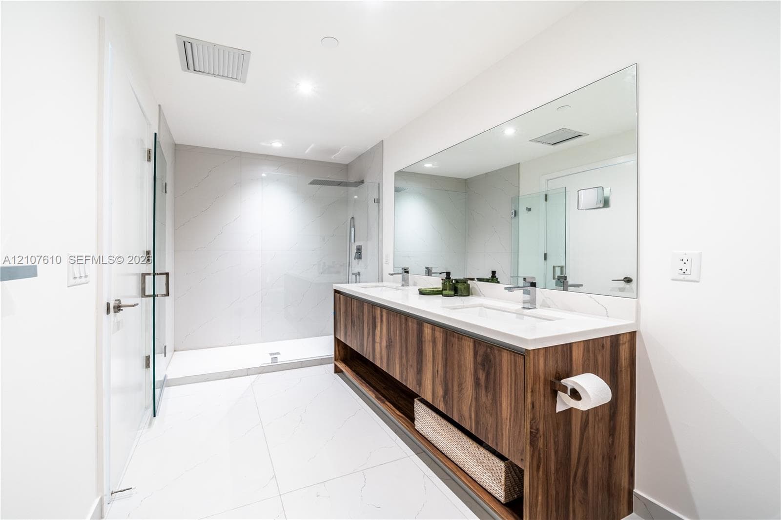 Dual-sink vanity with wood-finish cabinetry, stone countertop, large wall-mounted mirror, and walk-in shower featuring glass enclosure and stone tile surround