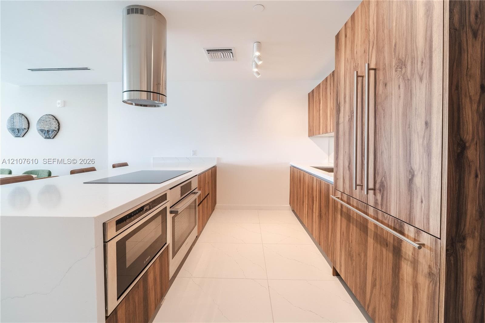 Kitchen featuring wood-finish cabinetry, white stone-surface countertops, integrated stainless steel appliances, a cylindrical ceiling-mounted range hood, and large-format tile flooring