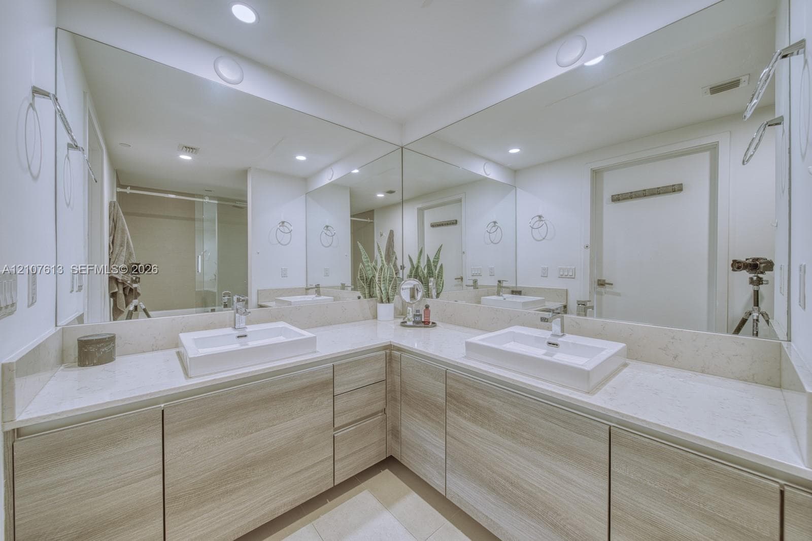 L-shaped wood-finish vanity with dual rectangular vessel sinks, light-toned stone countertops, full-width wall mirror, and integrated recessed lighting