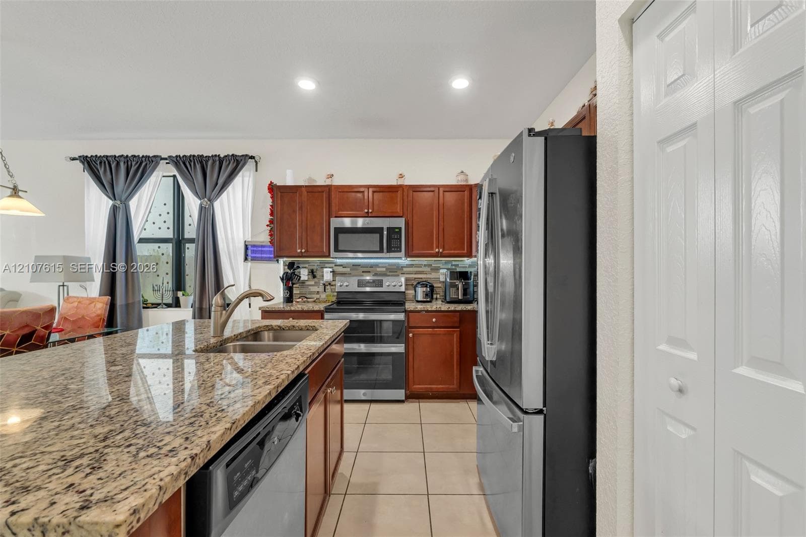 Kitchen featuring a spacious stone-surfaced island with an integrated sink, natural wood-finish cabinetry, tile flooring, and stainless steel appliances