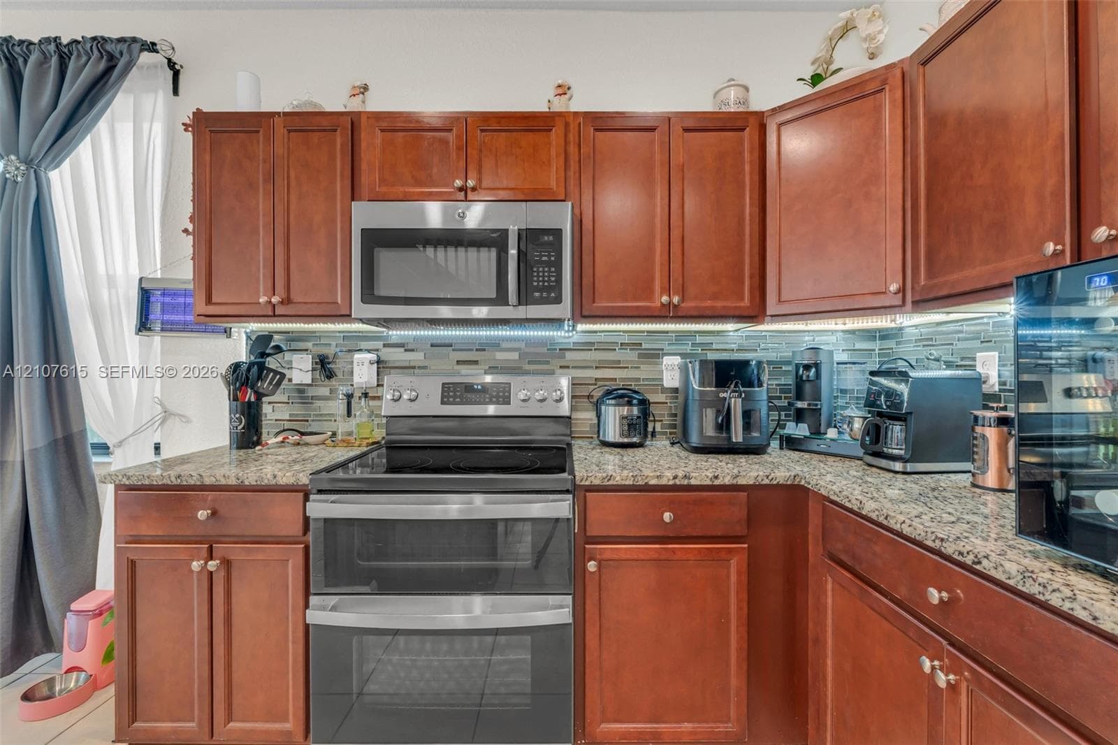 Kitchen featuring raised-panel wood cabinetry, speckled stone-finish countertops, stainless steel microwave, glass-top range with dual ovens, and a multi-toned horizontal tile backsplash